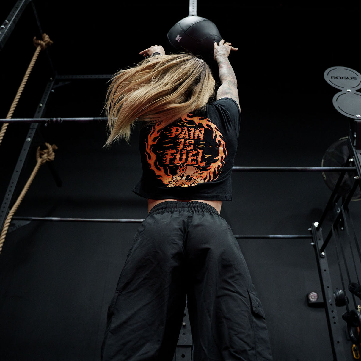 Female model doing a wall ball throw in a gym wearing black crop top with 'Pain Is Fuel' graphic